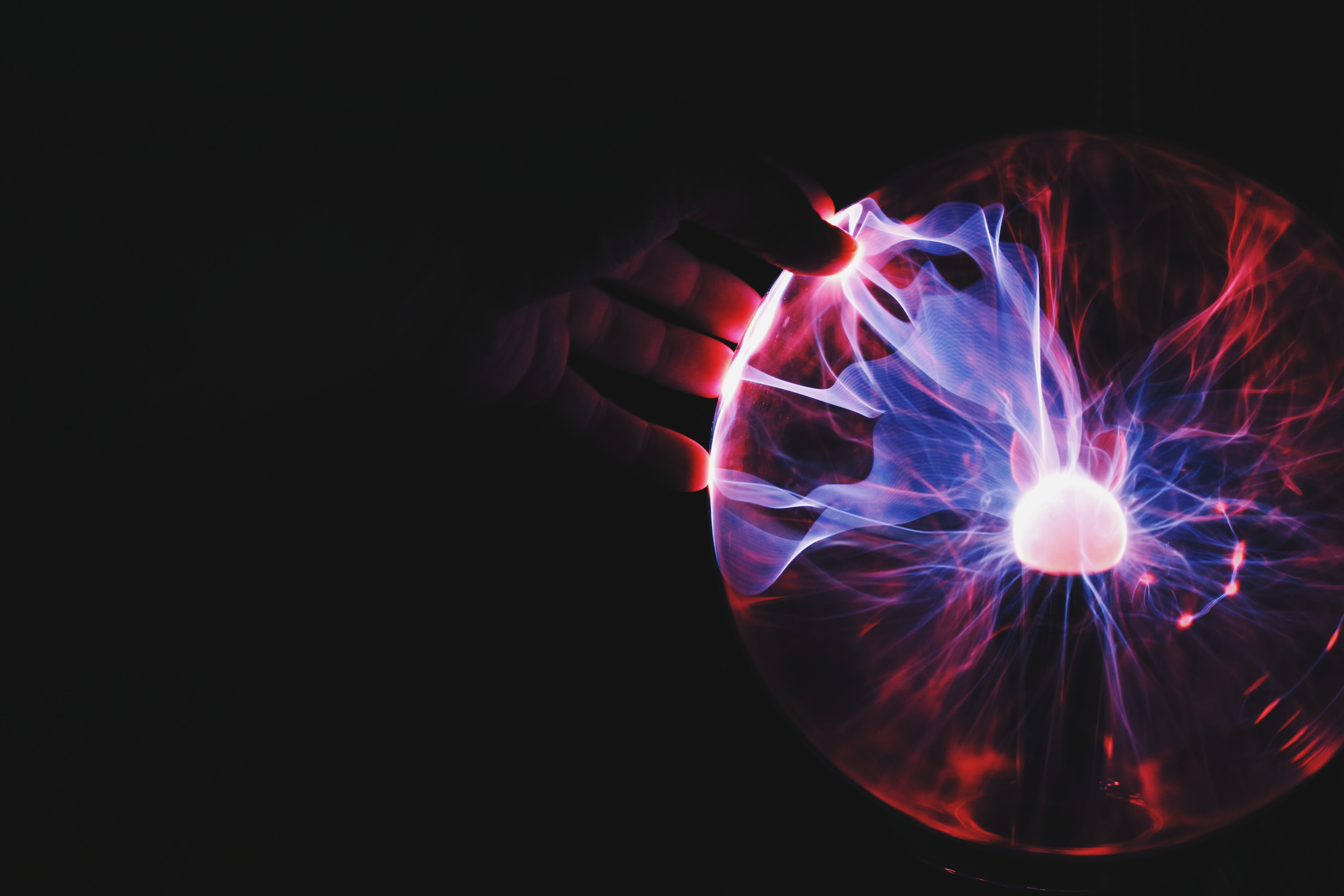 Mão toca esfera de plasma que emite raios vermelhos e azuis em fundo escuro, criando efeito luminoso e dinâmico.