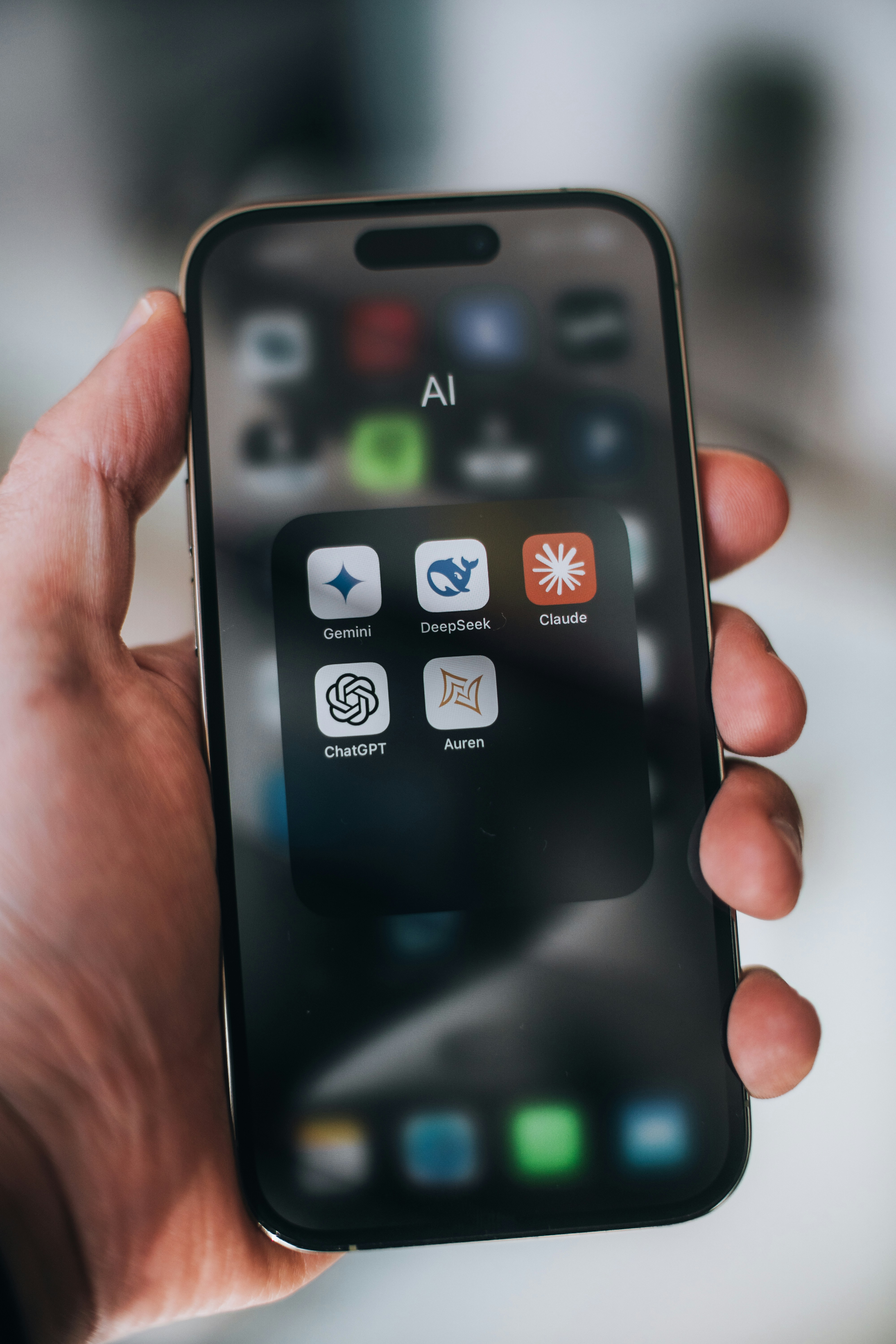 Mão segura um smartphone exibindo apps de IA como ChatGPT, Gemini, e Claude na tela. Fundo desfocado, ambiente neutro.