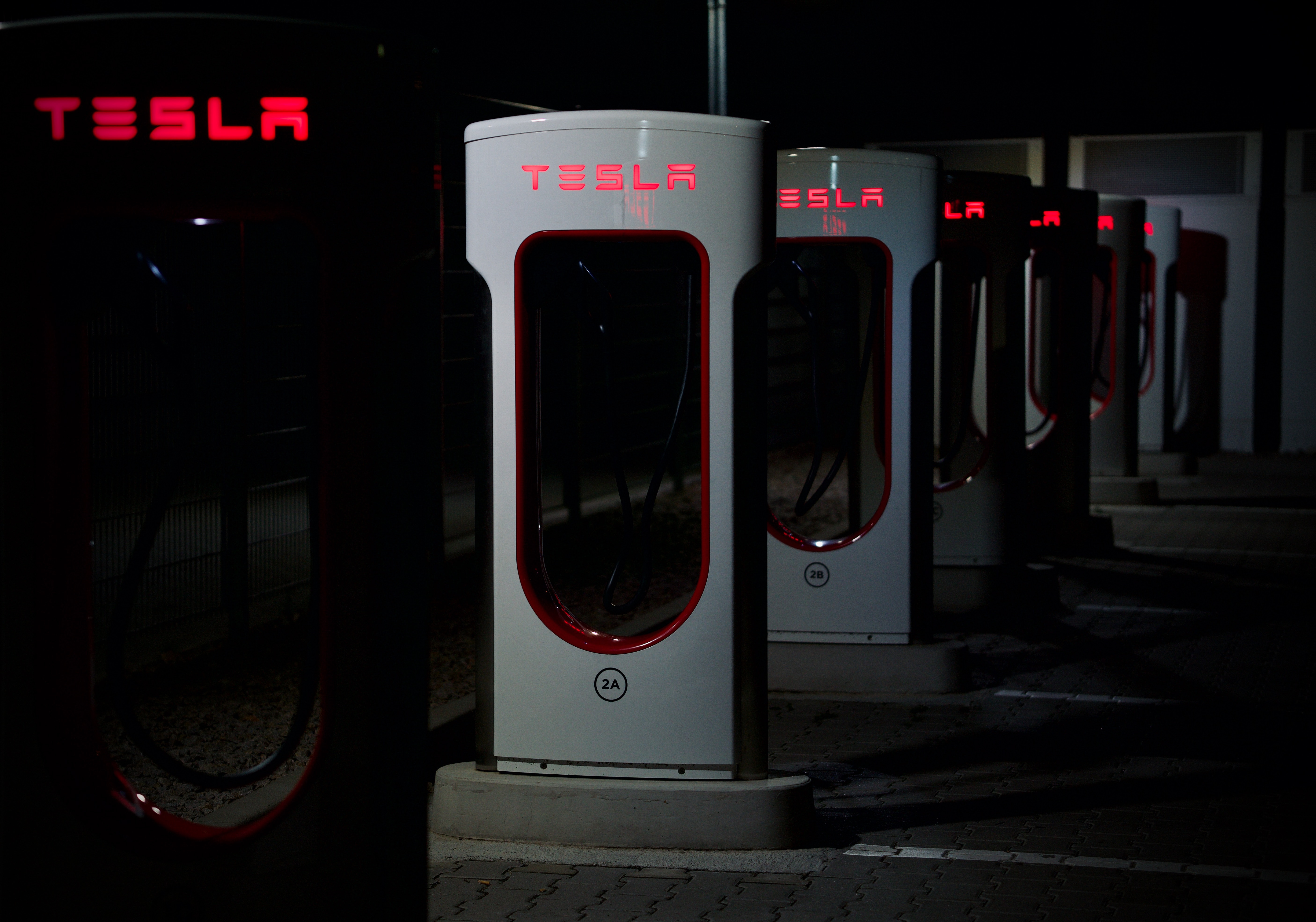 Estação Tesla Supercharger à noite com luzes vermelhas acesas. Cabos pendem, criando um clima tecnológico e moderno no escuro.